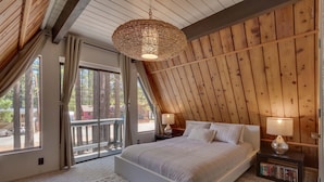 A bedroom with a large bed, wooden walls, a hanging light fixture, a bookshelf, and a view through a glass door.