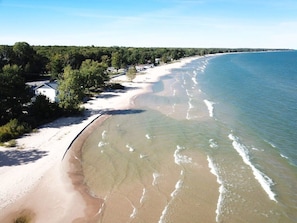 Una playa costera con orilla arenosa, cielo despejado y una zona boscosa al fondo.