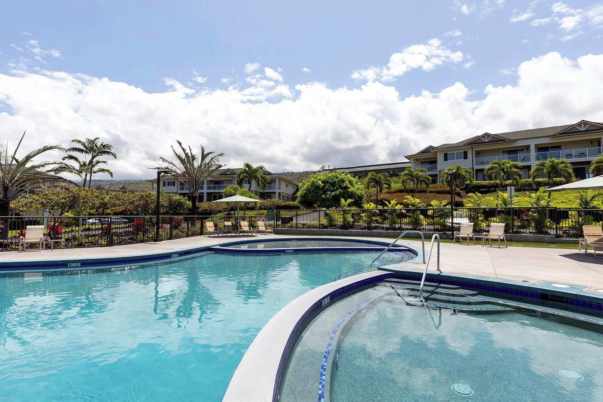 A swimming pool with lounge chairs and palm trees, surrounded by a fence and a building with multiple balconies.