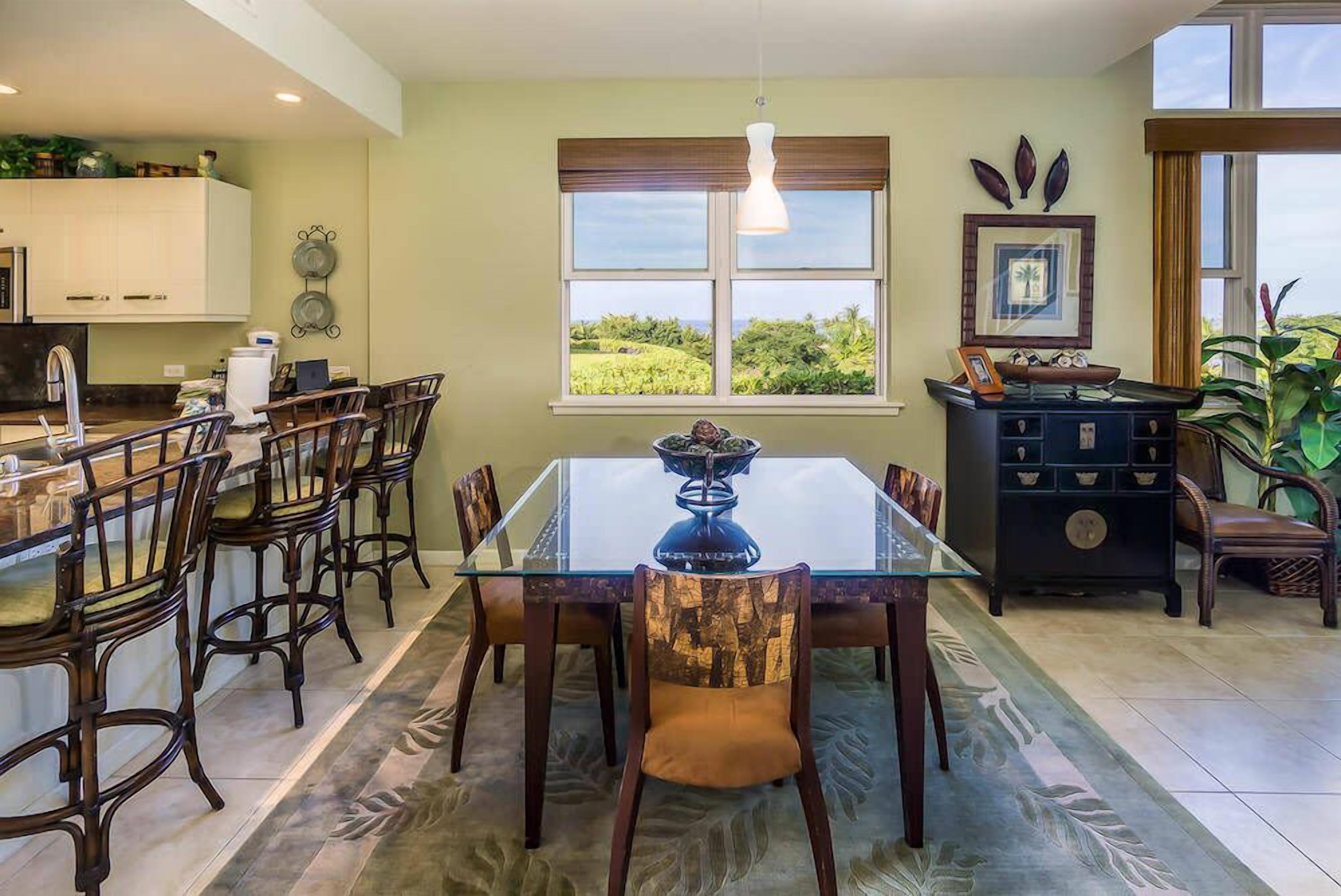 A dining area with a glass table and wooden chairs, a kitchen with white cabinets, and a window with a view of greenery.