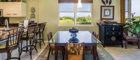 A dining area with a glass table and wooden chairs, a kitchen with white cabinets, and a window with a view of greenery.