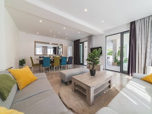 Una sala de estar moderna con un sofá, un reposapiés y una mesita de centro. Hay una zona de comedor con sillas y una mesa, y un ventanal con cortinas.