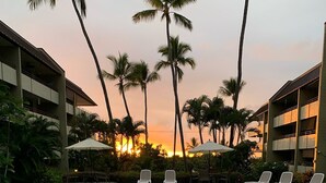 A swimming pool with lounge chairs and umbrellas, surrounded by palm trees and a building with balconies. - White Sands Village #201 (Kailua-Kona)