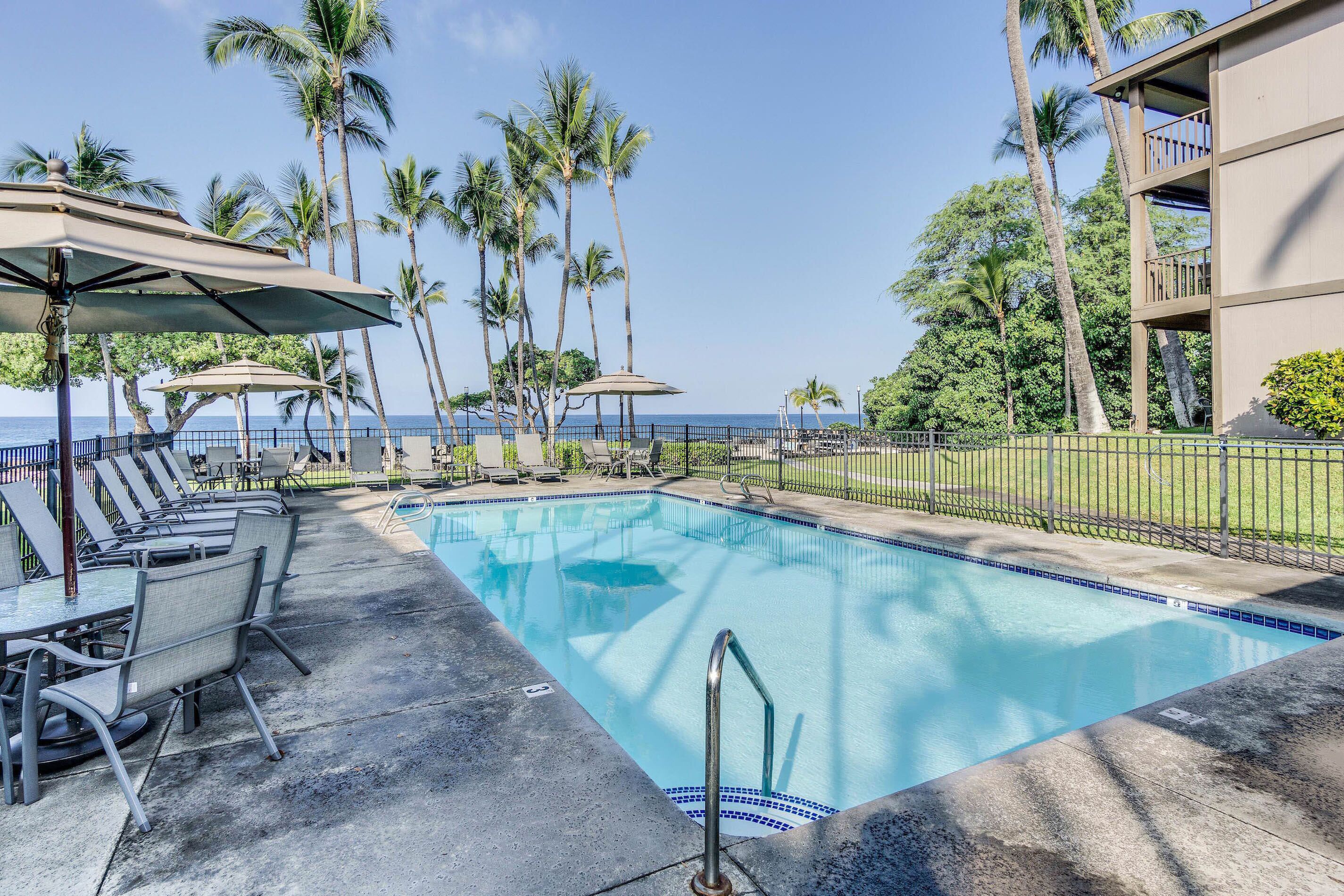 Ein Swimmingpool mit Liegestühlen und Sonnenschirmen, umgeben von Palmen und Blick aufs Meer.
