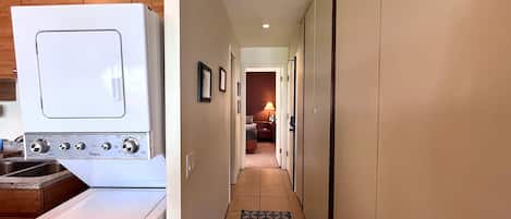A laundry area with a white washer and dryer, a hallway with a patterned rug, and a door leading to a bedroom.