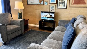 A living room with two grey sofas, a TV on a stand, a lamp, and various framed pictures on the wall. - Sea Village #1303 Ocean Front! (Kailua-Kona)