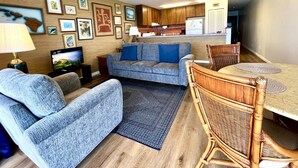 A living room with a grey sofa, a blue armchair, a dining table with a wicker chair, and a kitchen in the background. - Sea Village #1303 Ocean Front! (Kailua-Kona)