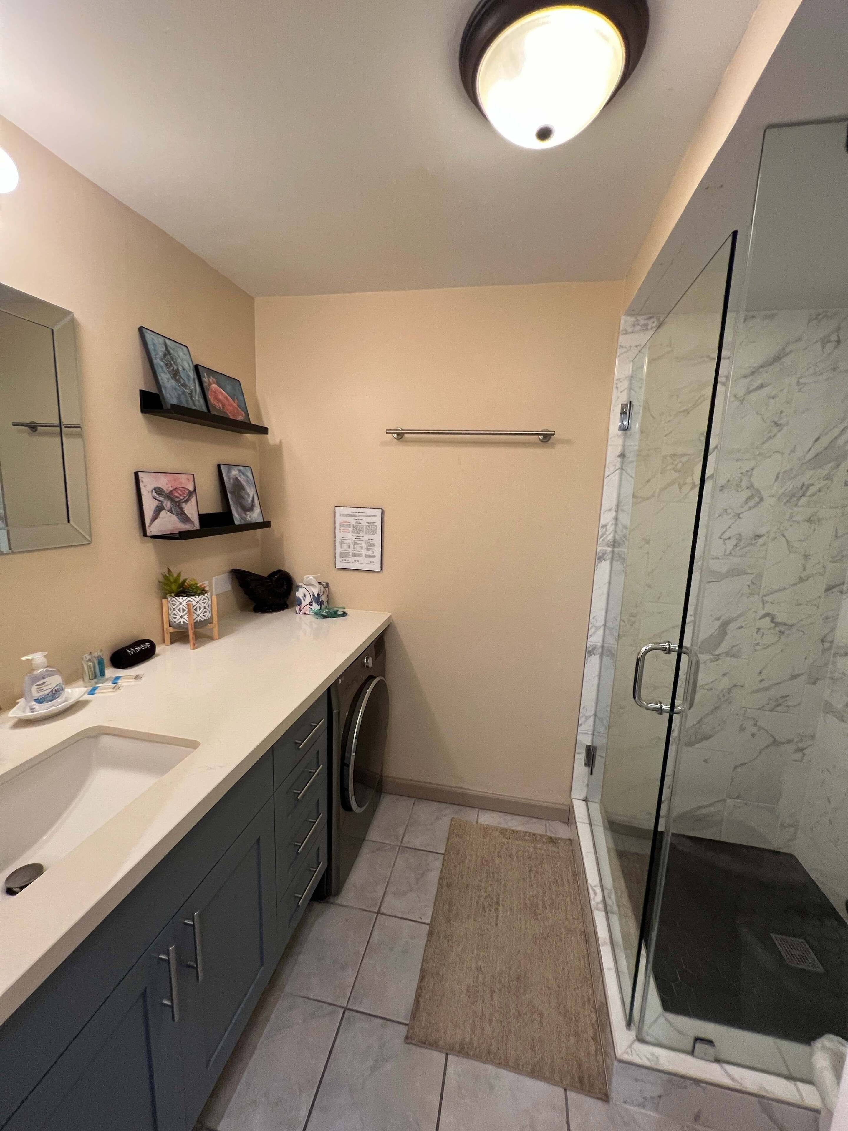 A bathroom with a sink, mirror, shelves with artwork, a towel rack, a shower with glass doors, and a small rug.