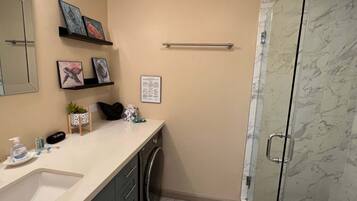 A bathroom with a sink, mirror, shelves with artwork, a towel rack, a shower with glass doors, and a small rug.