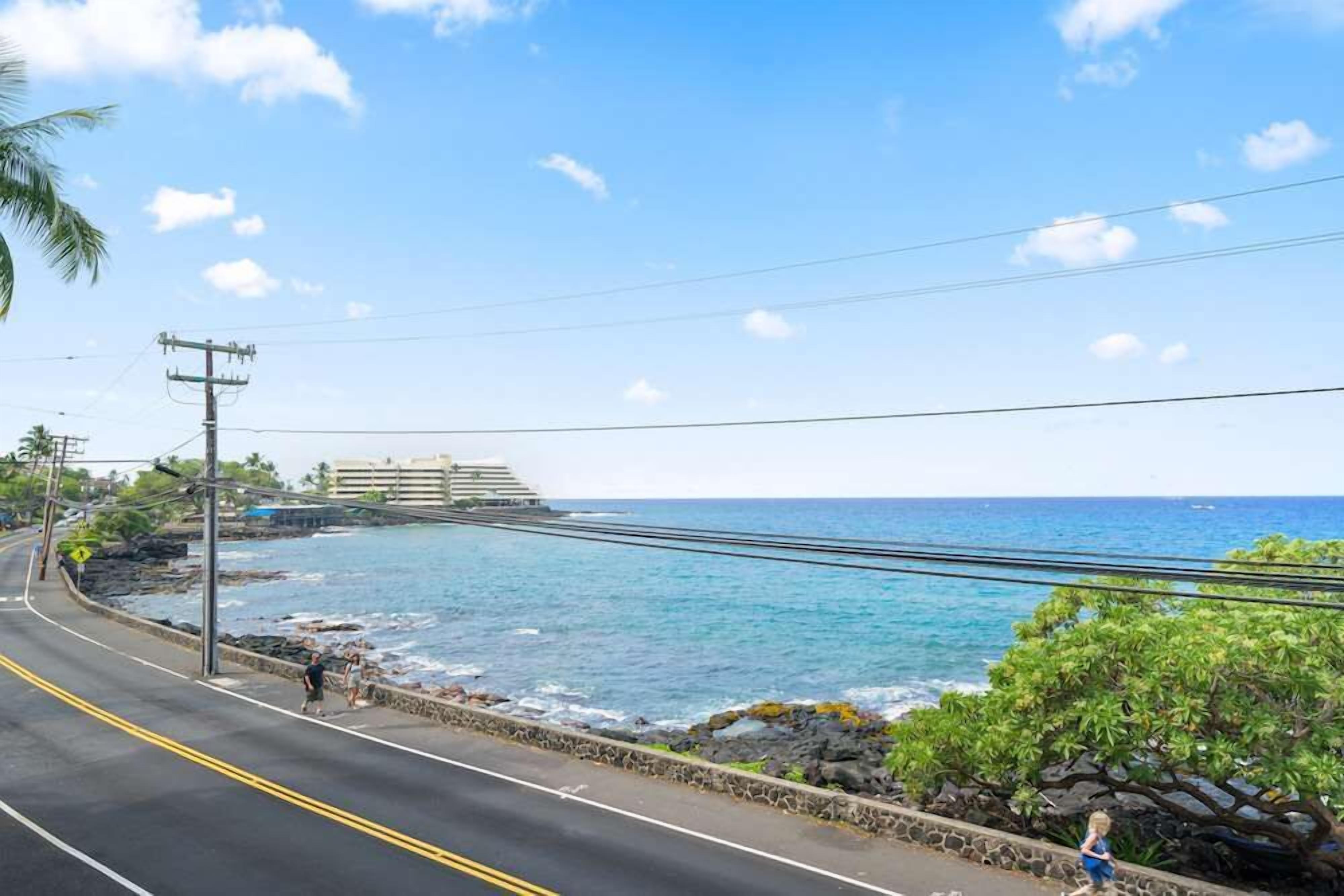 A coastal road with a view of the ocean, a pedestrian path, and a building in the background.