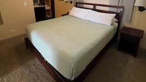 Ein Schlafzimmer mit einem Bett, einem Deckenventilator, einem Nachttisch, einer Lampe, einem Fenster mit Vorhängen und einem an der Wand befestigten Regal.