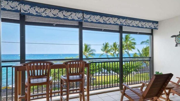 Ein Balkon mit Holzstühlen und einem Tisch, der Blick auf einen tropischen Strand mit Palmen und das Meer bietet.