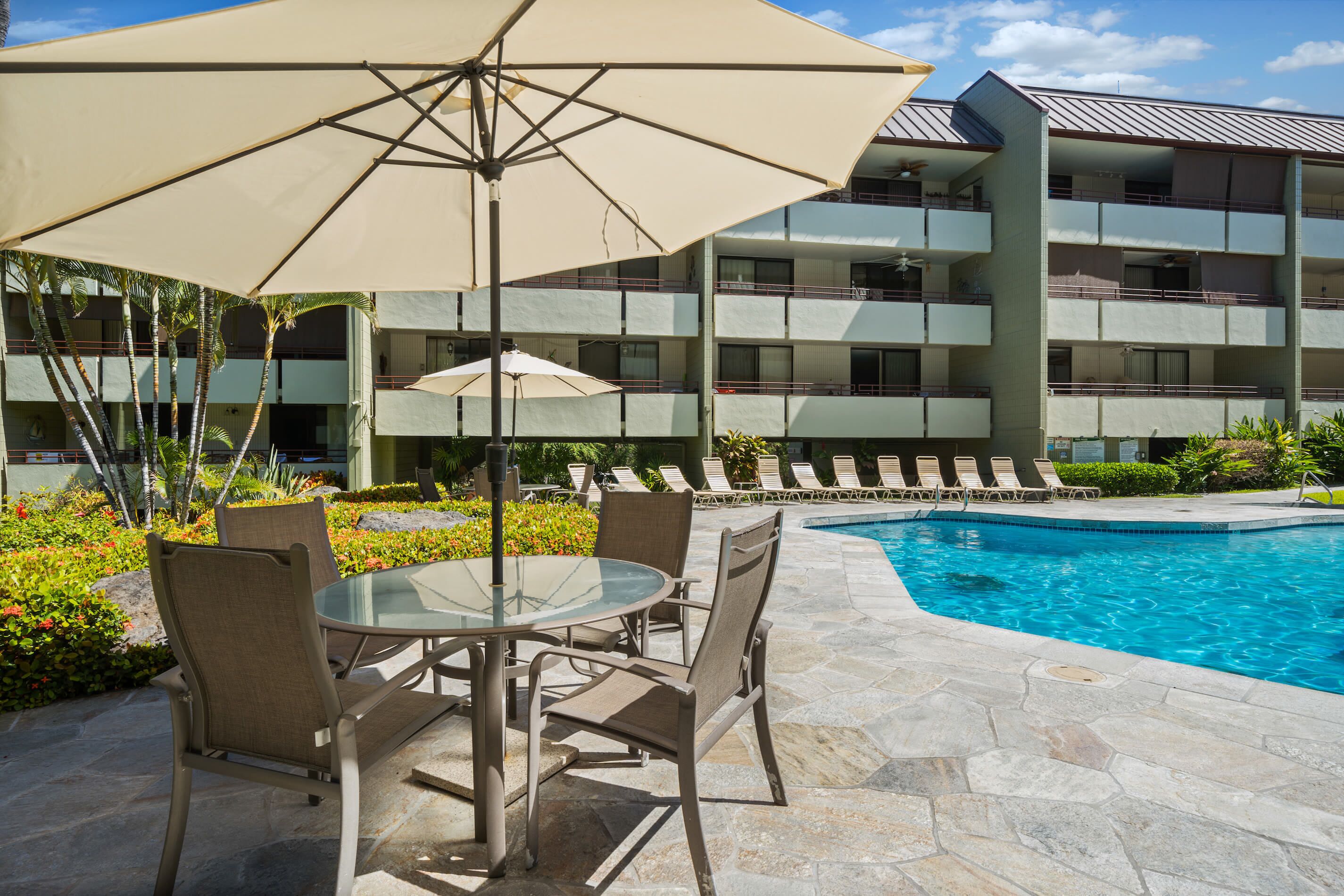 A poolside area with a glass table and chairs, a large umbrella, and a building with balconies in the background.