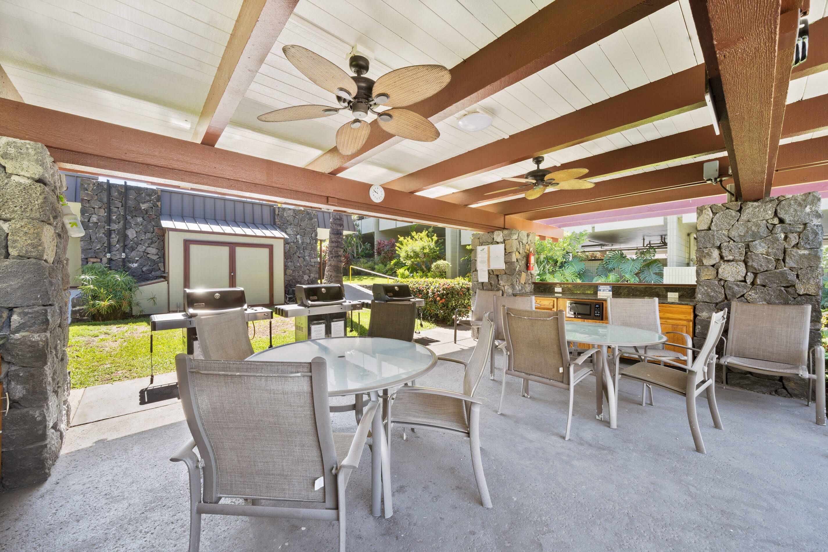 An outdoor patio with a round glass table and four chairs, a ceiling fan, and a stone wall.