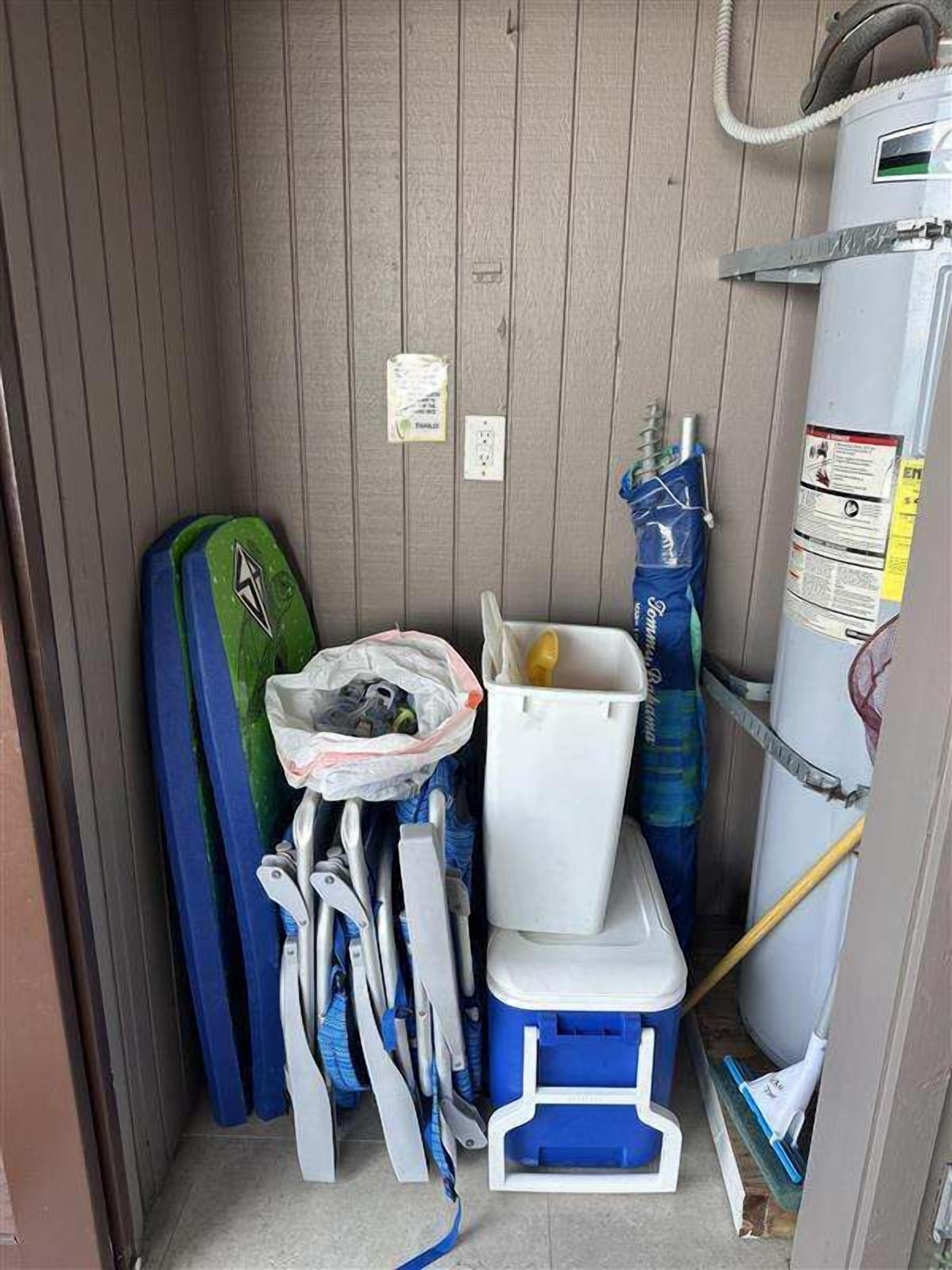 Une petite pièce étroite aux murs en bois, avec un radiateur blanc, une boîte de rangement bleue et une planche de surf verte et bleue appuyée contre le mur.
