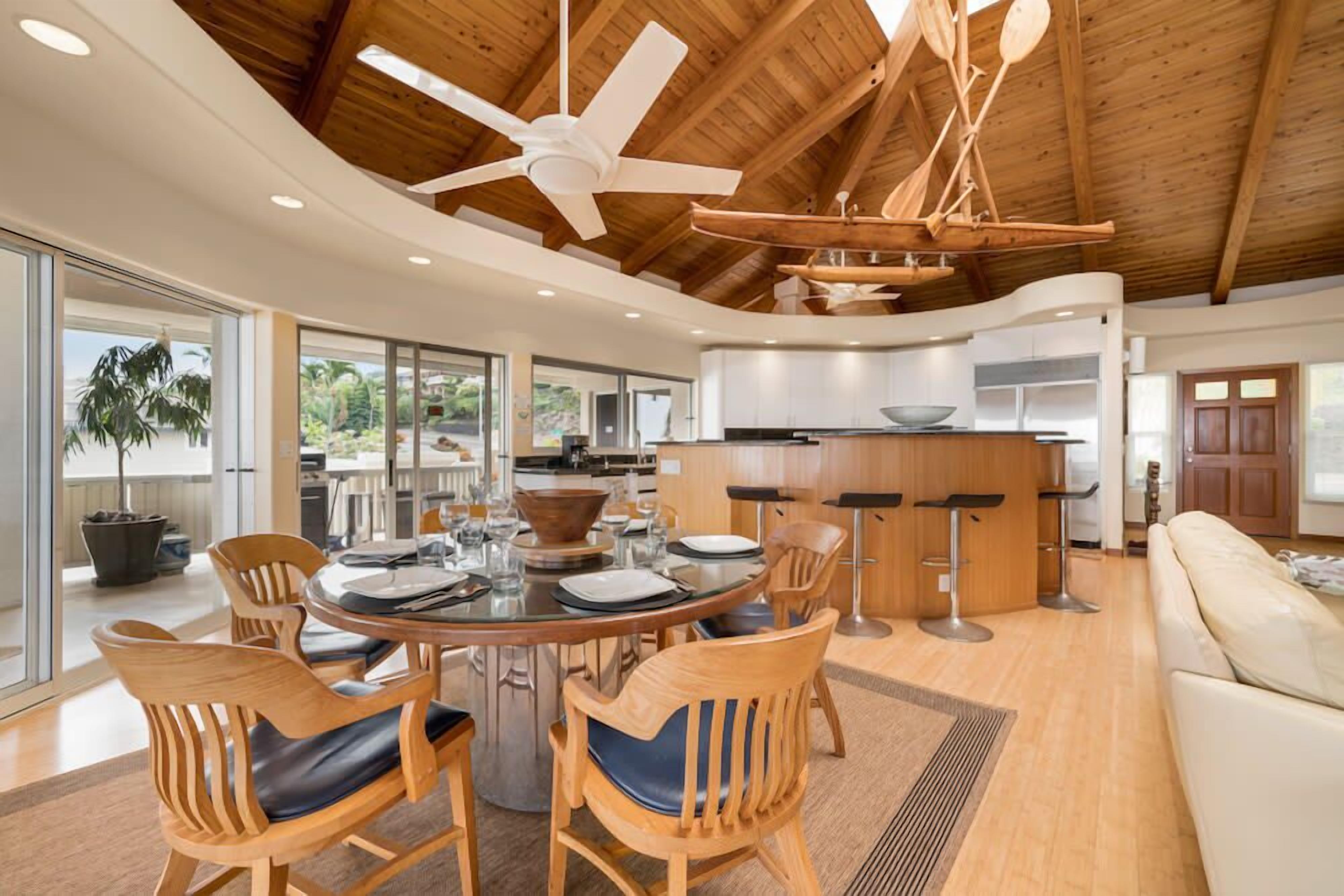 A spacious living area with a wooden ceiling, a round dining table surrounded by chairs, a kitchen area with a bar, and a large window offering a view of the outdoors.