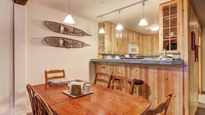 A wooden dining table with chairs, a kitchen area with wooden cabinets, and pendant lights.