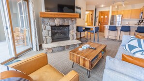 A modern living room with a stone fireplace, a wooden coffee table, and a sofa. There is a kitchen area with wooden cabinets and a refrigerator.