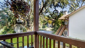 A wooden deck with a glass table and a chair, overlooking a garden with a pond and a house with a staircase.