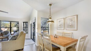 A dining area with a wooden table and white chairs, a hanging light fixture, and framed artwork on the wall.