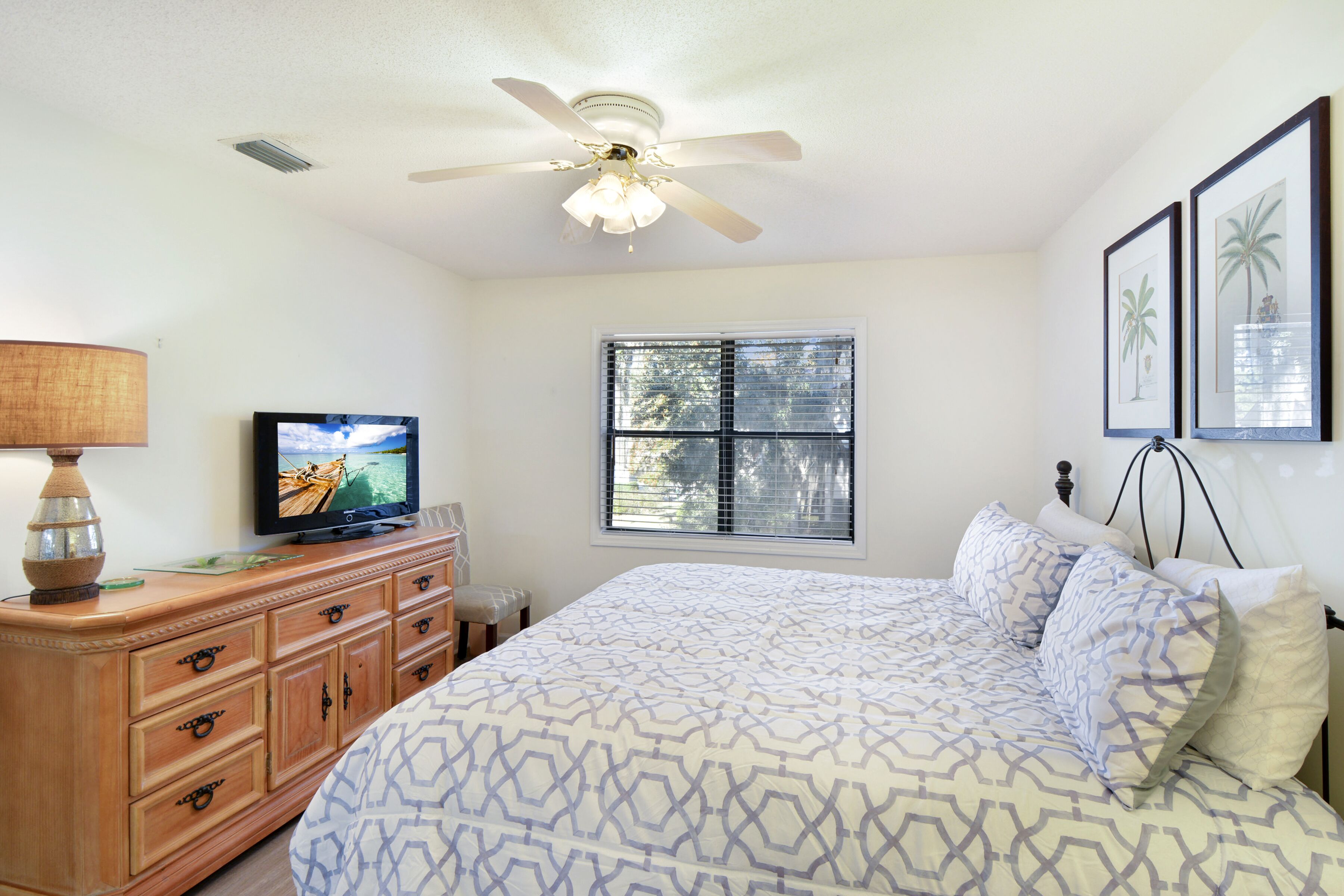 Une chambre à coucher avec un lit, un téléviseur posé sur une commode en bois, un ventilateur de plafond et une fenêtre avec des stores.
