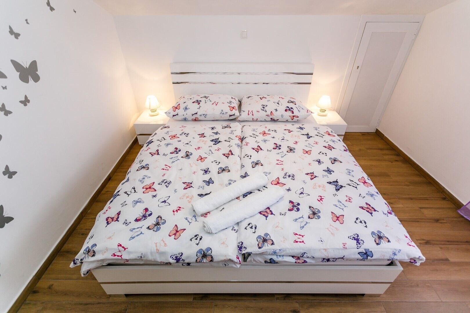 Uma cama com colcha estampada com flores e borboletas, duas luminárias laterais e um cabeceira branca.
