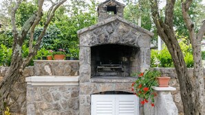 Un horno de pizza de piedra al aire libre con chimenea, rodeado de árboles y plantas en macetas.