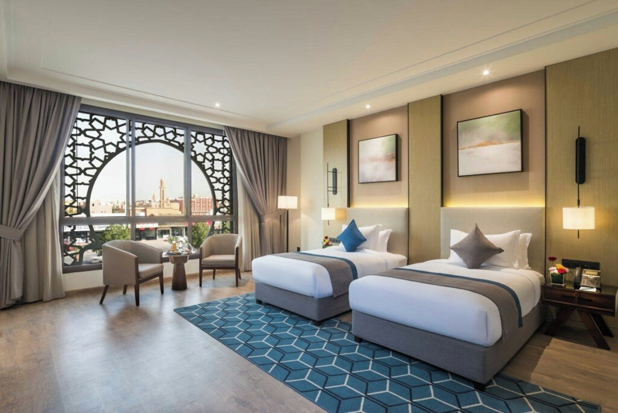 A hotel room with two beds, a seating area with chairs, a view through a decorative window, and a patterned rug.