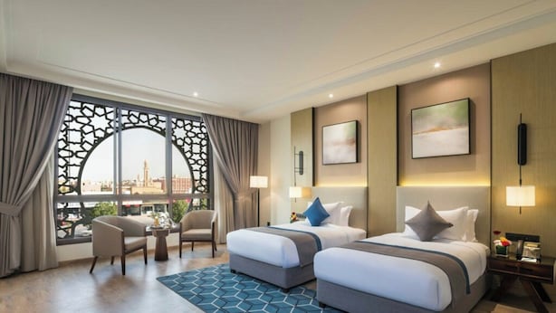 A hotel room with two beds, a seating area with chairs, a view through a decorative window, and a patterned rug. - Braira Al Yarmouk Hotel (Riyadh)