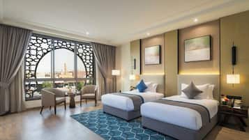 A hotel room with two beds, a seating area with chairs, a view through a decorative window, and a patterned rug.