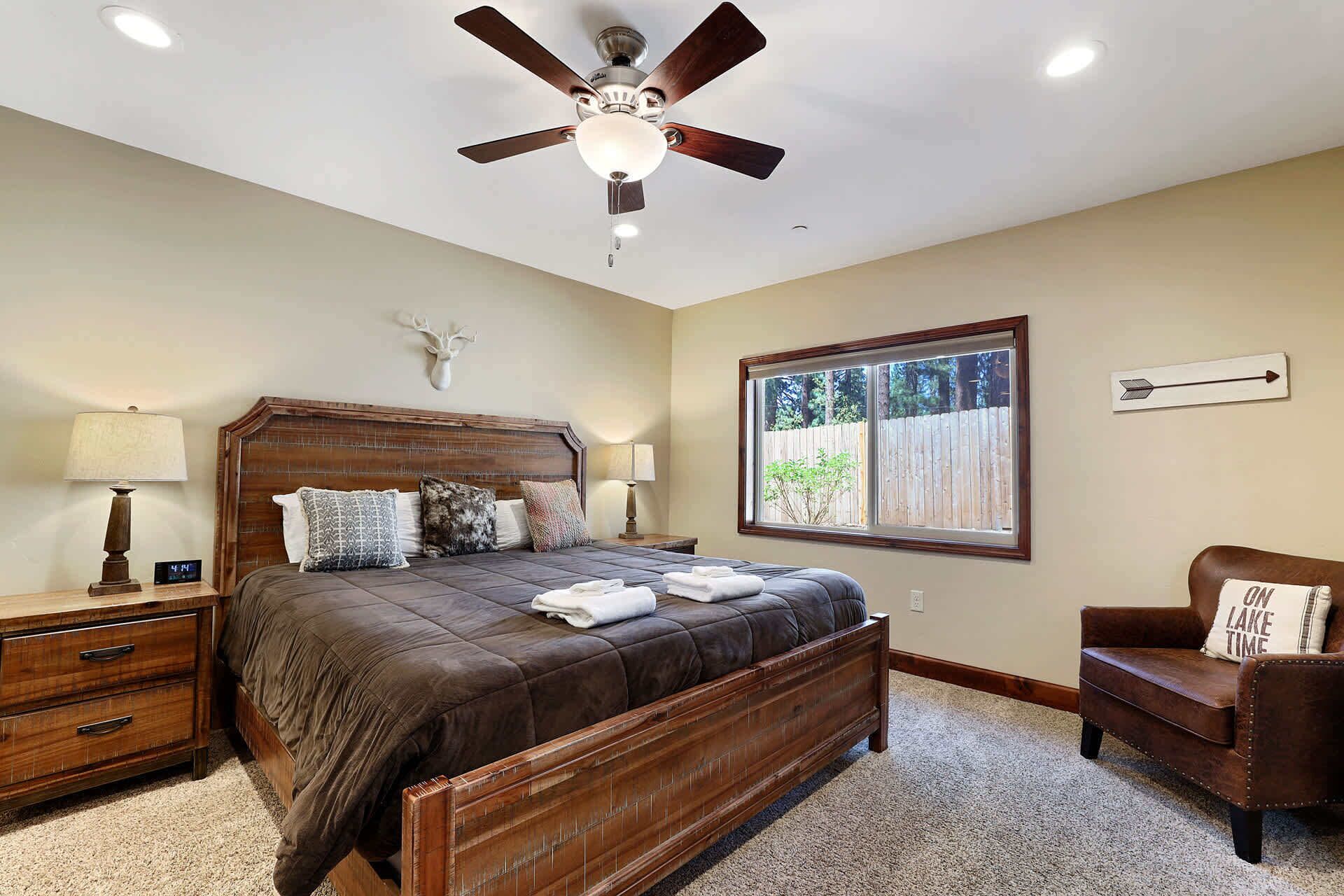 A bedroom with a wooden bed, a ceiling fan, a nightstand with a lamp, a wooden dresser, a window with a view of trees, and a wall-mounted deer head decoration.