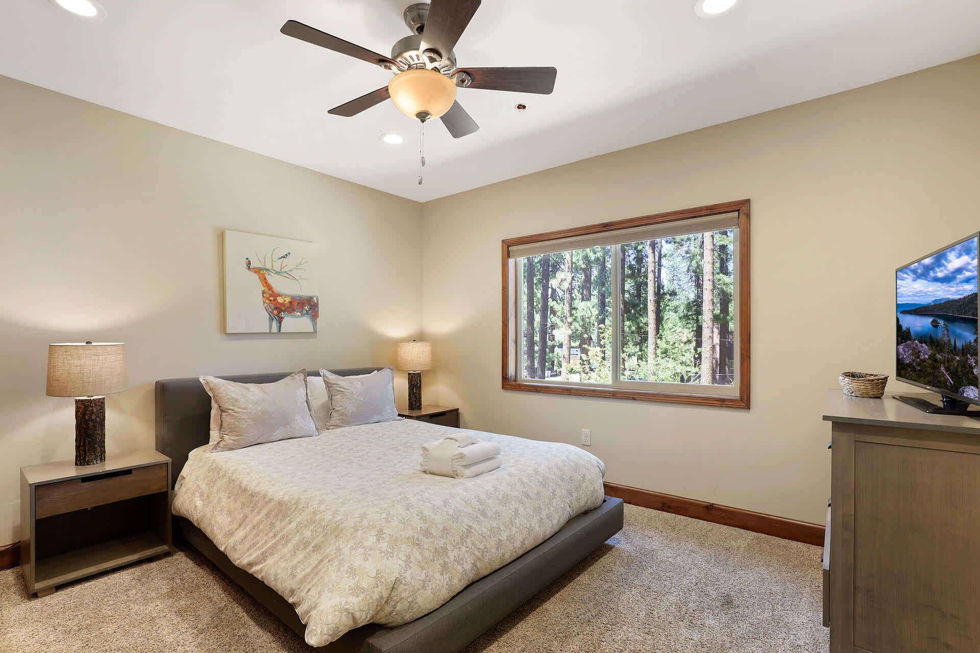 Un dormitorio con cama, ventilador de techo, ventana con vista al bosque y televisor.