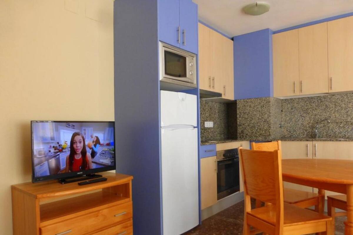 Cocina compacta con microondas, horno y refrigerador. Hay una mesa de madera y una silla, y un televisor sobre un mueble de madera.