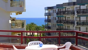 Un balcon avec des chaises et une table en plastique blanc, donnant sur un immeuble doté de nombreux balcons et offrant une vue sur la mer.