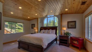 Une chambre à coucher avec un grand lit, deux tables de chevet avec des lampes, un poêle à bois rouge et une grande fenêtre donnant sur la forêt.
