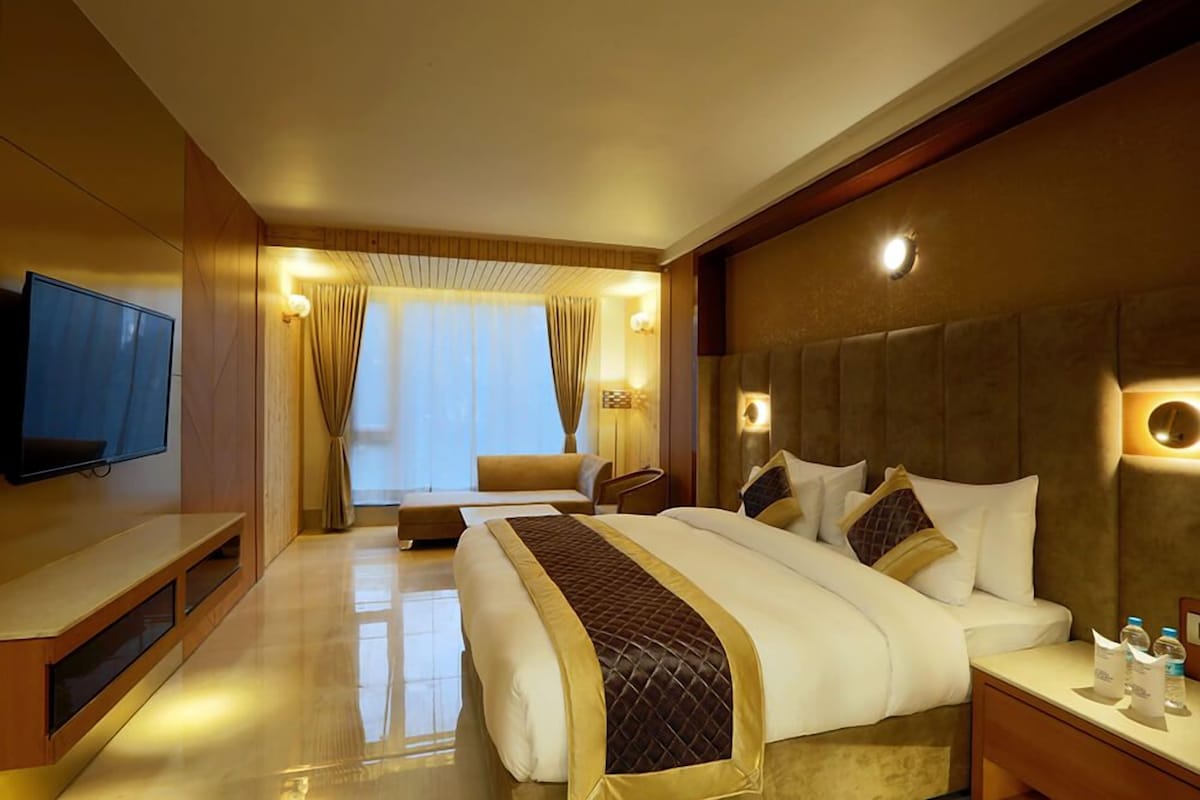 A hotel room with a large bed, a TV, a sofa, and a desk with a lamp.