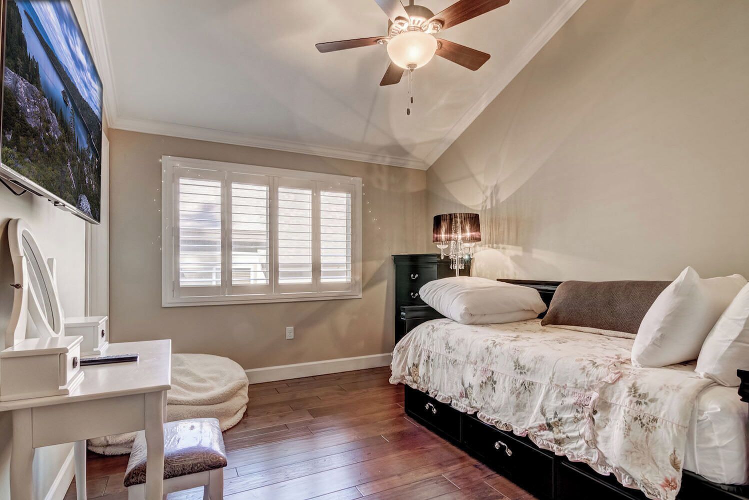 A bedroom with a bed, a desk, a chair, a vanity, a ceiling fan, and a window with blinds.