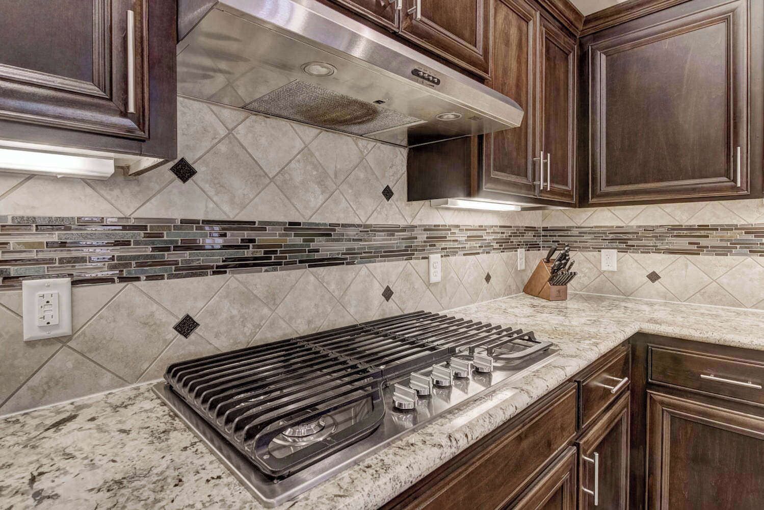 A kitchen with dark wood cabinets, a stainless steel range hood, a tiled backsplash, and a granite countertop.