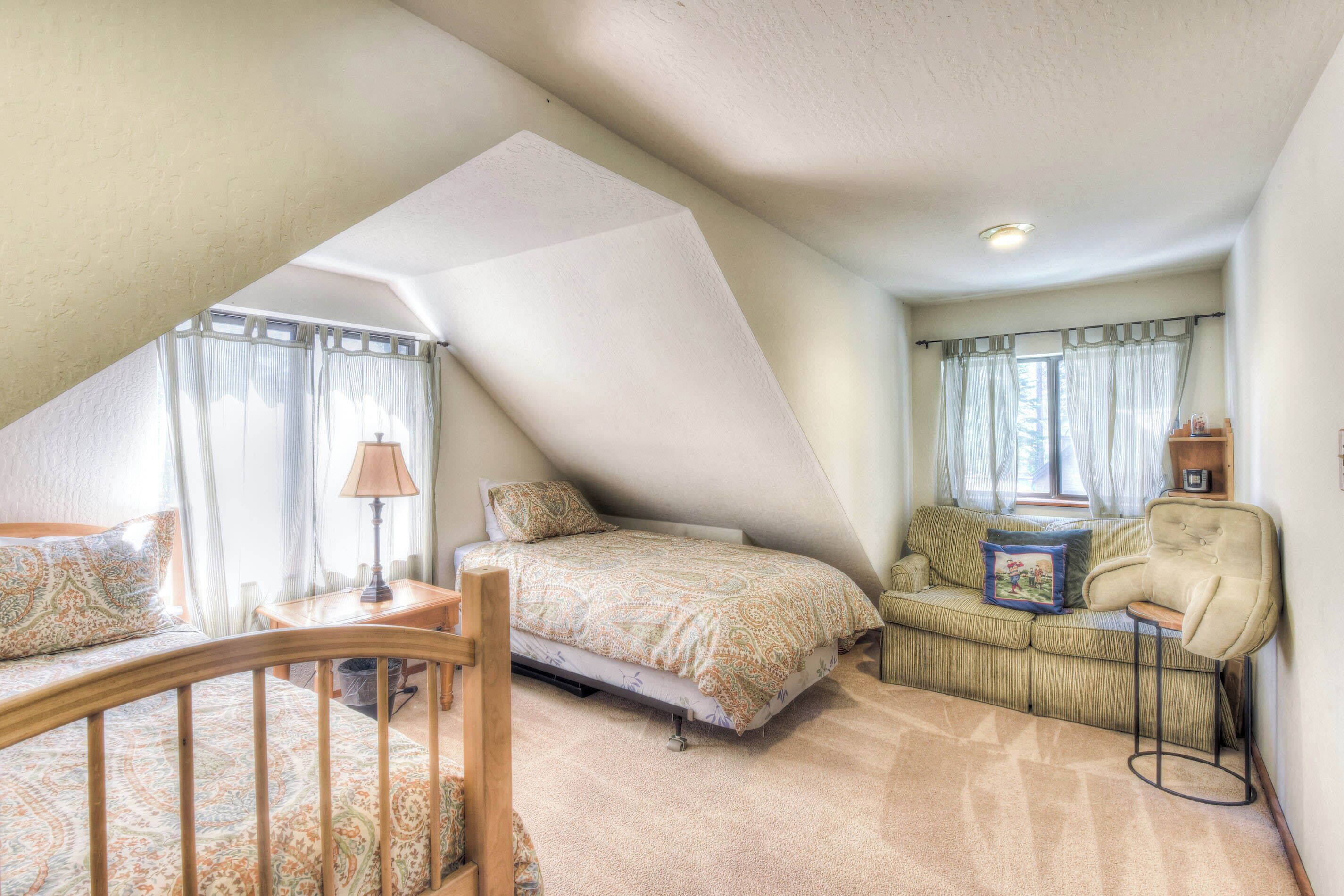 A cozy attic bedroom with a sloped ceiling, a bed with patterned bedding, a small table with a lamp, a sofa with cushions, and a chair.