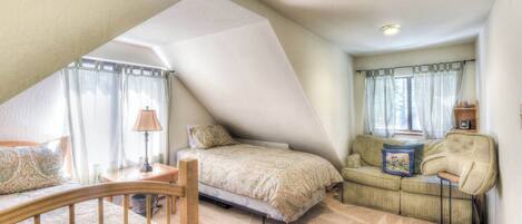 A cozy attic bedroom with a sloped ceiling, a bed with patterned bedding, a small table with a lamp, a sofa with cushions, and a chair.
