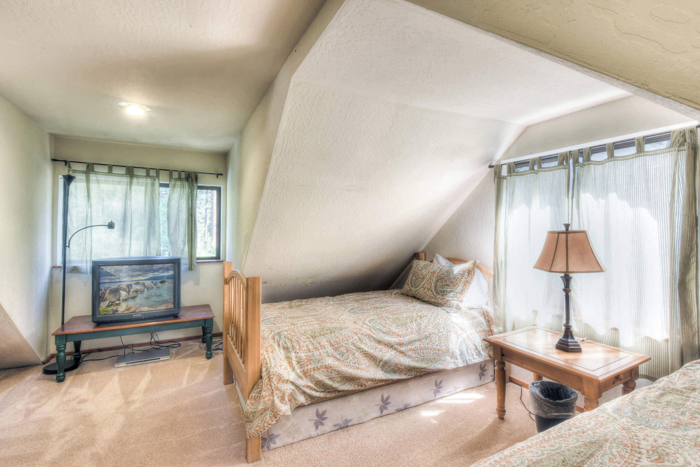 Ein Schlafzimmer mit schrägen Wänden, einem Bett mit gemusterten Betttextilien, einem Nachttisch mit Lampe, einem Fernseher auf einem Schrank und einem Fenster mit Vorhängen.