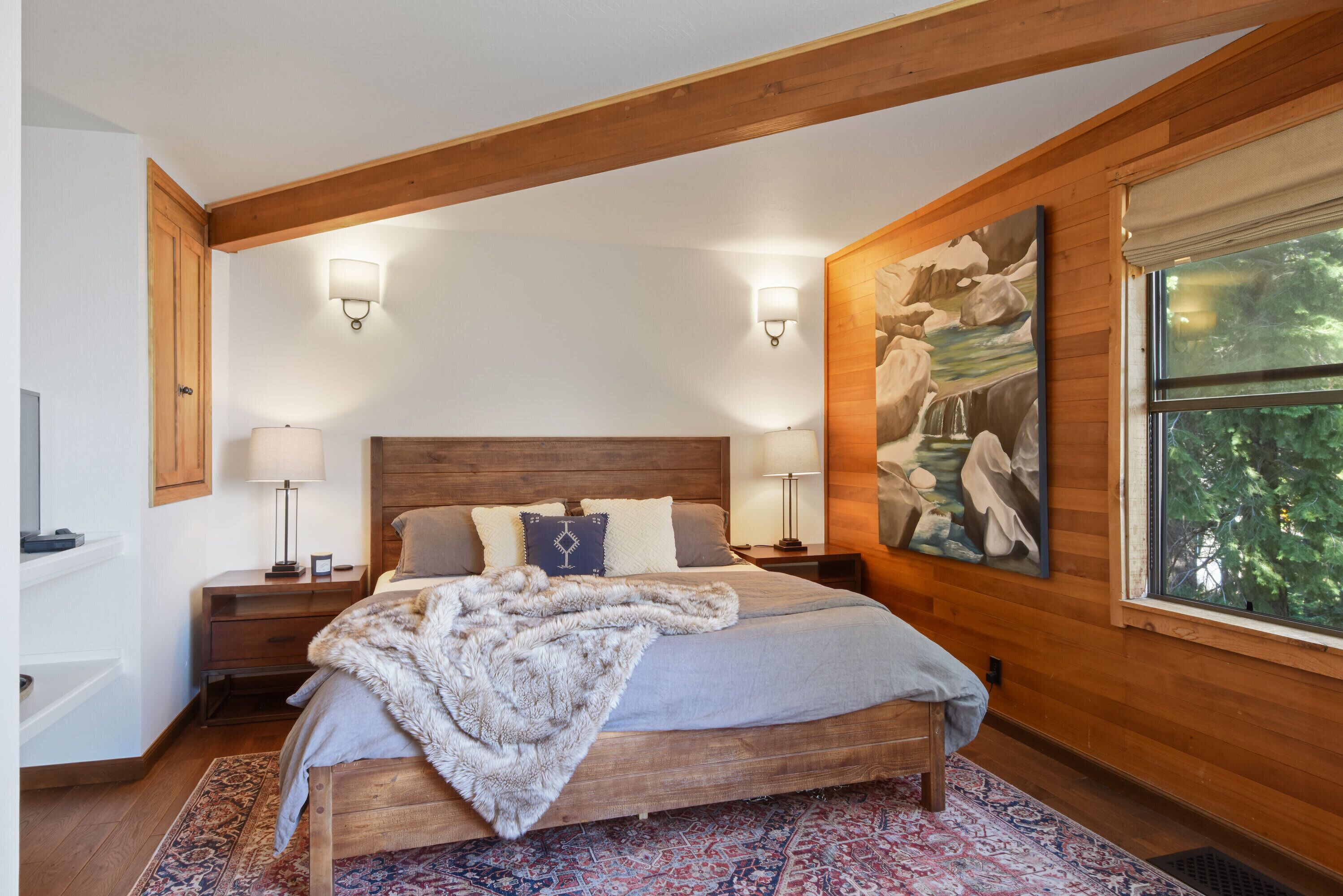 A bedroom with a wooden headboard, two bedside lamps, a large painting, and a window with a view of trees.