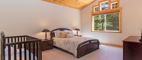 A bedroom with a wooden ceiling, a bed with a patterned bedspread, two bedside lamps, a crib, and a window with a view of trees.