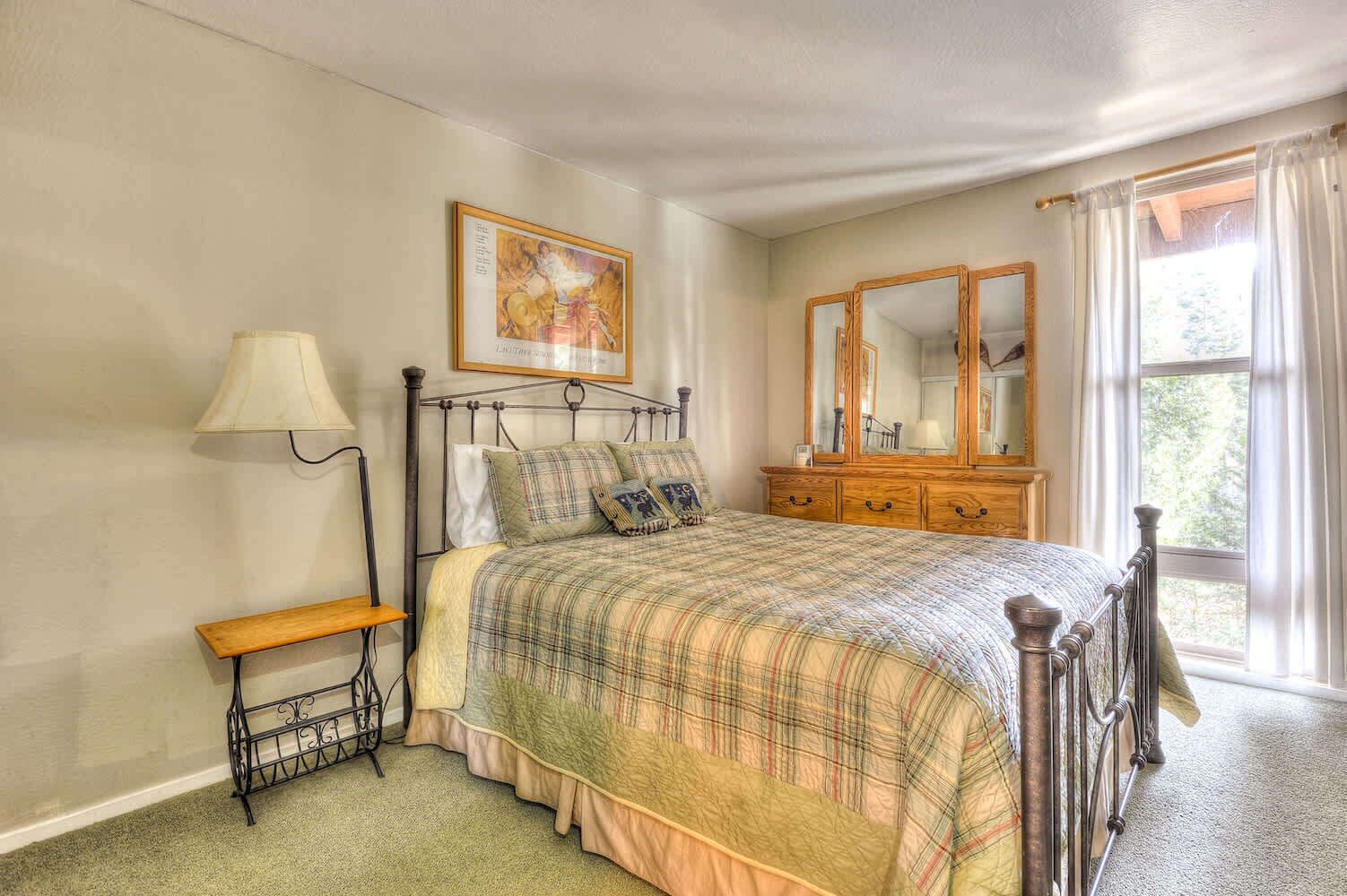 A bedroom with a bed, a bedside table, a lamp, a dresser, a mirror, and a window with curtains.