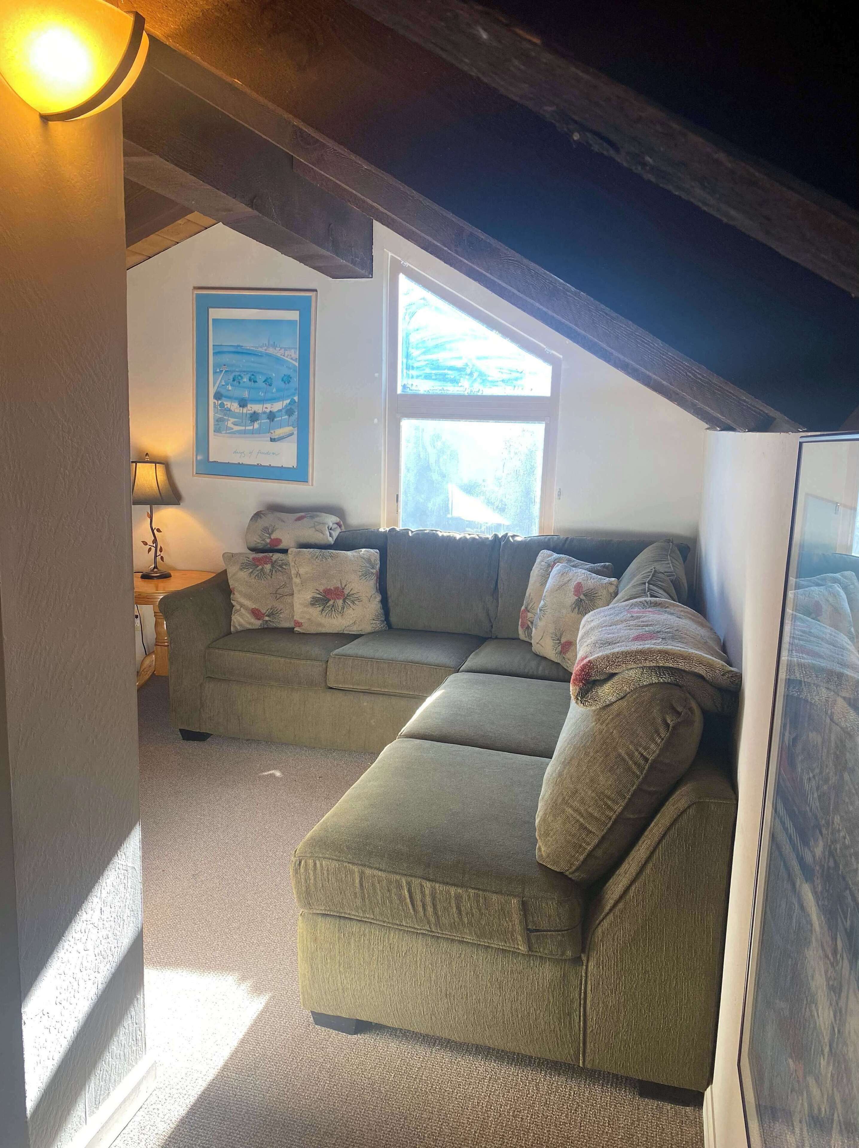 A cozy attic room with a green sectional sofa, a framed landscape painting, and a window providing natural light.