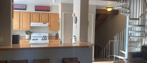 A modern kitchen with wooden cabinets, a central island with stools, a stove, and a sign that reads "LATTE." A staircase leads to a living area with a couch and a coffee table.