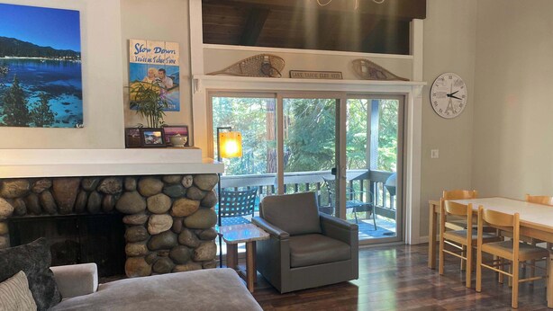 A cozy living room with a stone fireplace, a sofa, a dining table with chairs, and a view of the outdoors through large sliding glass doors. - Lake Forest Villa Patio w/ BBQ (Tahoe City)