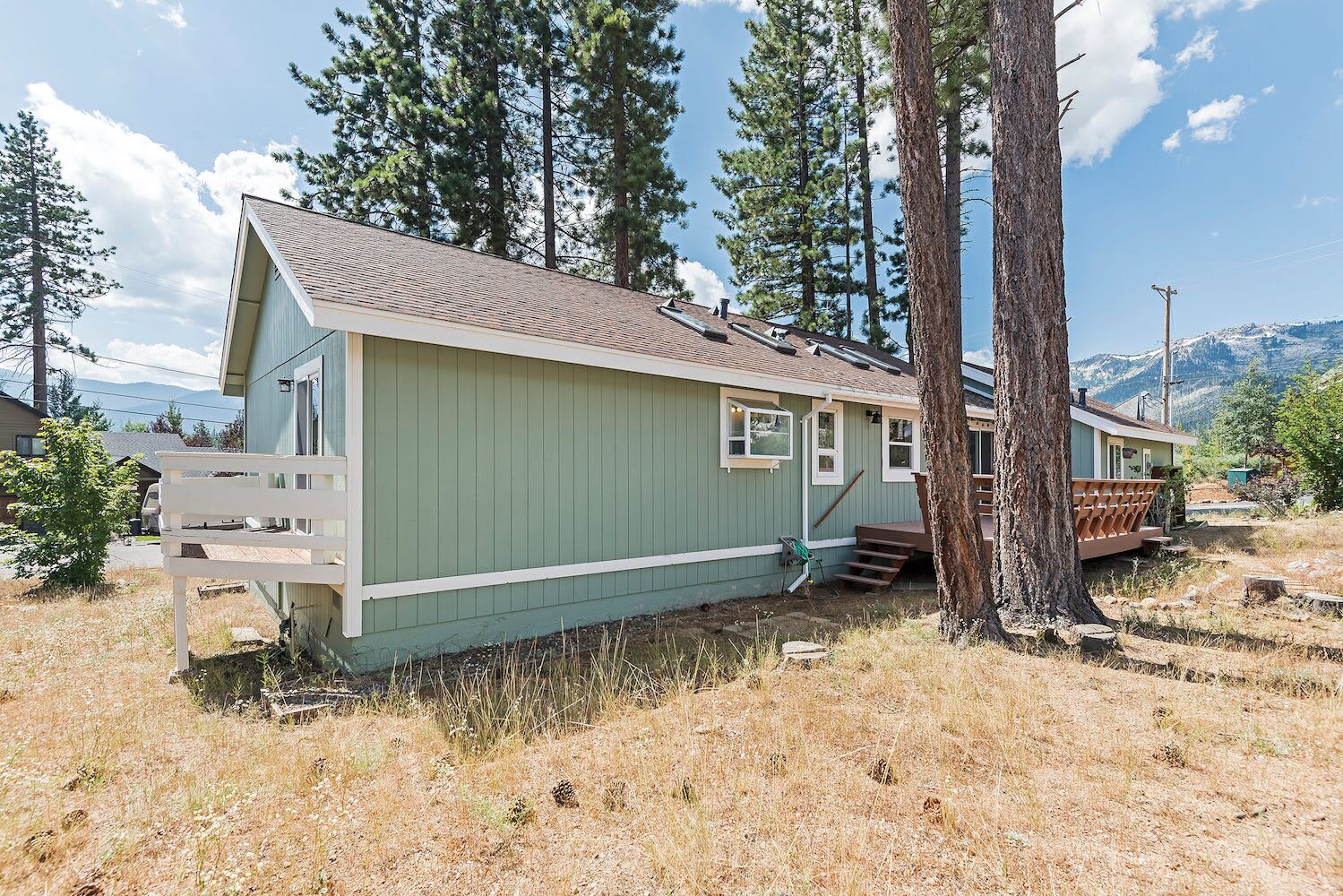 A single-story, green mobile home with a porch and a small deck, surrounded by trees and a mountainous backdrop.