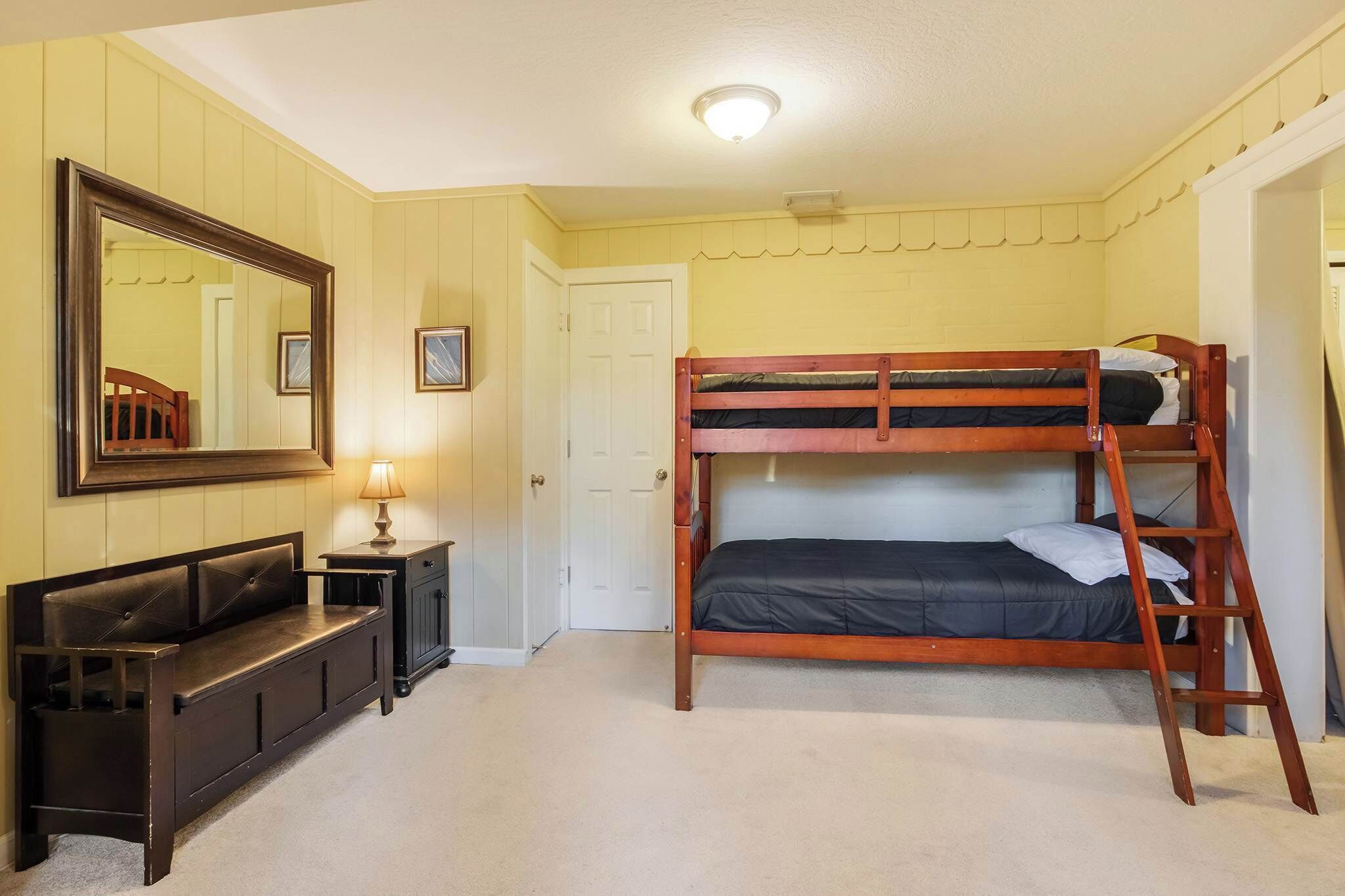 Een stapelbed met een ladder, een bank, een bureau, een spiegel en een deur in een kamer met gele wanden.