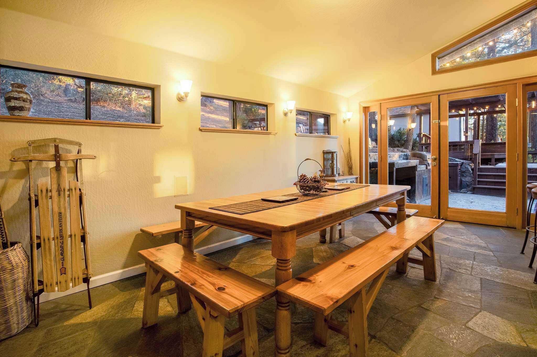 A wooden dining table with benches, a stone floor, and a view through glass doors to an outdoor area.